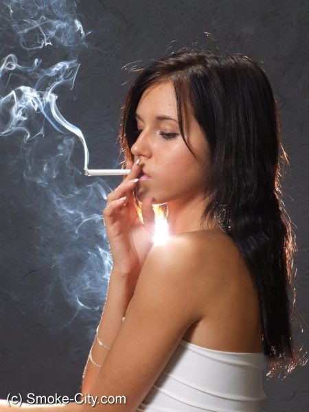 Молодая брюнетка курит сигарету, одетая в обтягивающее белое платье и туфли на каблуках